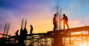 Contingent workers standing on scaffolding at construction site. Dramatic sunset in background.